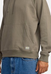 Oliwkowy hoodie wykonany z miękkiej bawełny, z kieszenią kangurką, długimi rękawami, prążkowanymi mankietami oraz małą metką z logo blisko hemu.
