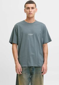 Graues T-Shirt mit kurzen Ärmeln und Rundhalsausschnitt, vorne mit einem kleinen weißen „JJ Studios“-Logo. Weiches Baumwollmaterial, lockere Passform.