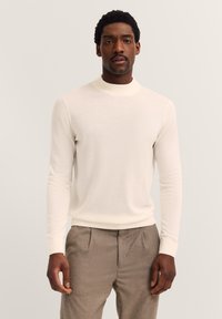 Hellecremiger, langärmeliger Pullover mit hohem Kragen und gerippten Bündchen, aus weichem Material. Kombiniert mit hellgrauen Hosen.