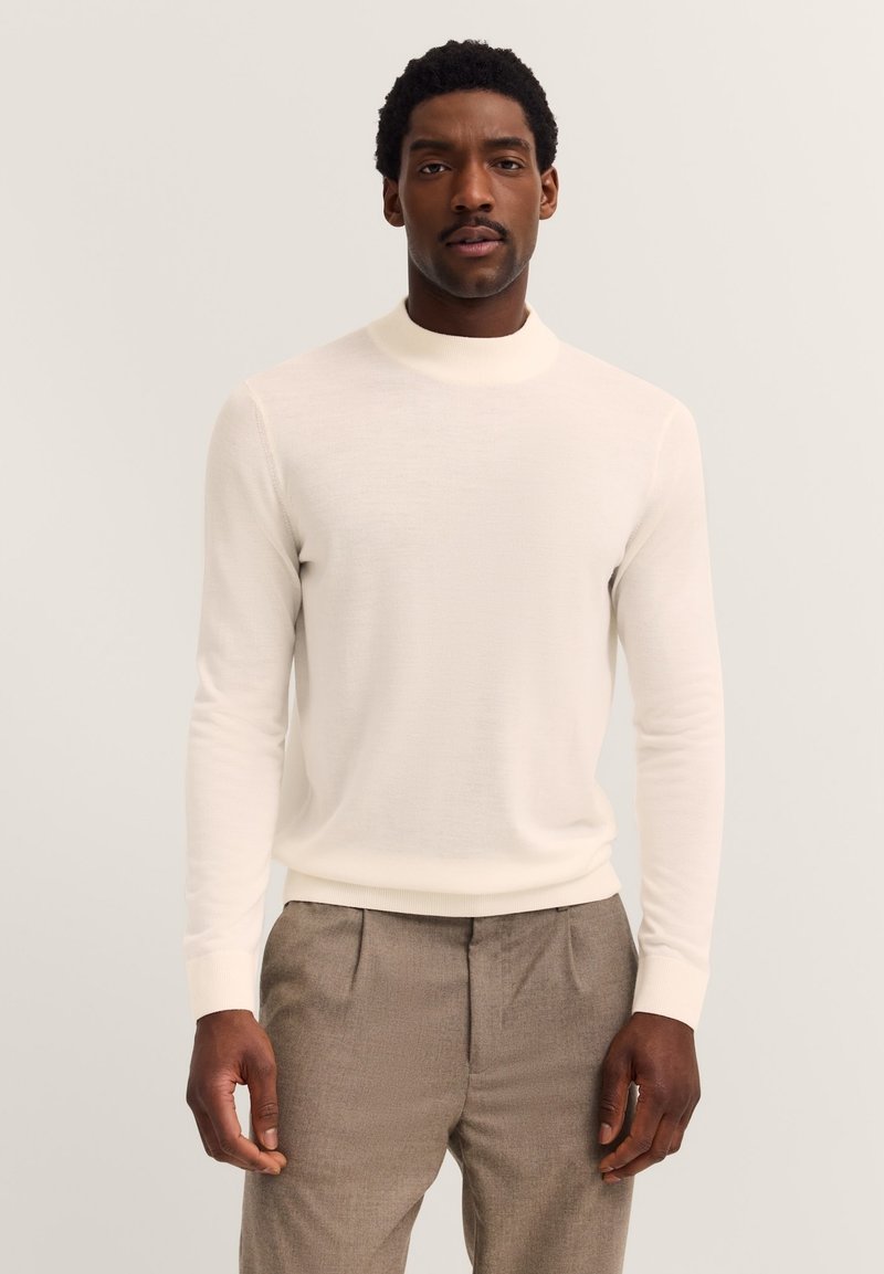 Hellecremiger, langärmeliger Pullover mit hohem Kragen und gerippten Bündchen, aus weichem Material. Kombiniert mit hellgrauen Hosen.