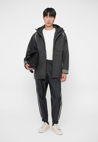 Svart vattentät jacka med grå detaljer, med två framfickor. Kombination med svarta träningsbyxor med vita ränder och beige sneakers.