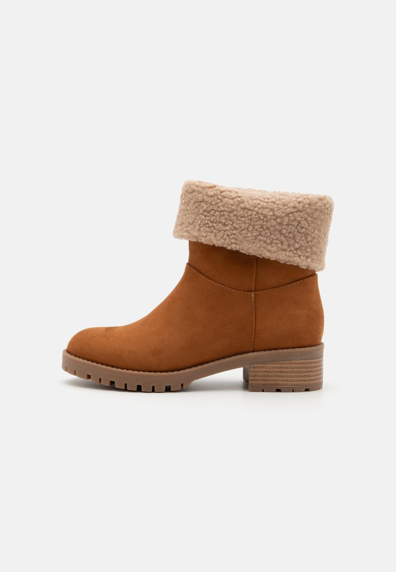 Bottine en suede marron avec un revers en fausse fourrure beige, texture lisse, bout rond et un talon empilé marron avec une semelle striée.