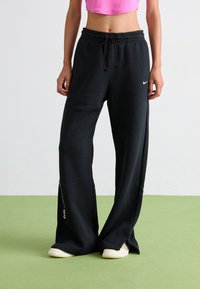 Schwarze Sweatpants aus weichem Material mit einem Kordelzugbund, weiten Beinen und seitlichen Schlitzdetails. Auffälliges Nike-Logo auf der Vorderseite.