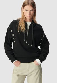 Felpa nera con dettagli metallici a occhiello sulle maniche, chiusura con zip e cordini testurizzati. Abbinata a pantaloni chiari.