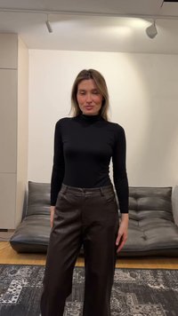 Γυναίκα με μακριά μαλλιά που φοράει μαύρο ζιβάγκο και σκούρο καφέ δερμάτινο παντελόνι, στέκεται σε ένα δωμάτιο με γκρίζο καναπέ και χαλί με σχέδια.