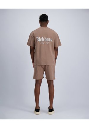 Mann trägt beige Shorts und passendes T-Shirt mit weißem Text "Blckbnns Design Dept" auf dem Rücken, steht vor einfachem grauem Hintergrund.