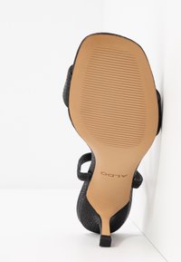 Sandale noire à talons hauts avec une tige texturée, semelle visible avec des rainures d'adhérence et le nom de la marque "ALDO" imprimé sur la semelle beige.