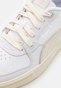 Zapatilla blanca con materiales de cuero y sintético. Presenta un lado texturizado, acentos en beige y cordones en crema. Punta perforada para ventilación.