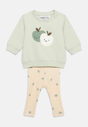 Haut vert clair à manches longues avec un motif de pommes souriantes vertes et blanches, associé à des leggings beiges ornés de motifs de pommes vertes éparpillées.