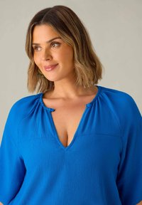 Blusa blu con texture, caratterizzata da un scollo a V, maniche a metà e un dettaglio arricciato al colletto. Tessuto morbido con motivi sottili.