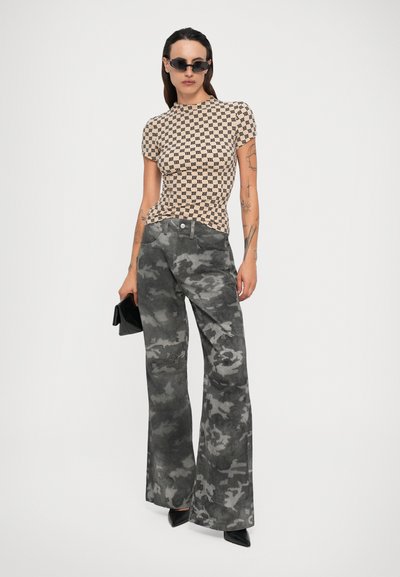Frau in beigem, gemustertem, kurzärmligem Oberteil, grauen Camouflage-weiten Hosen, schwarzen spitzen Schuhen, rechteckigen Sonnenbrille und einer schwarzen Clutch.