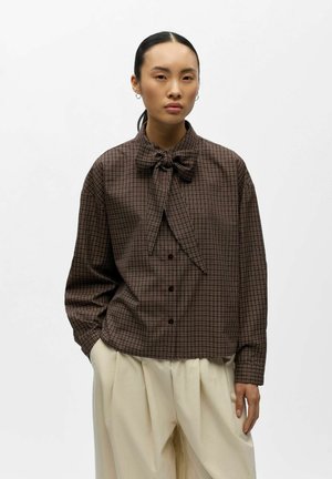 Femme portant une chemise à carreaux marron boutonnée avec un nœud papillon et un pantalon crème à jambes larges, debout les mains dans les poches sur fond blanc.