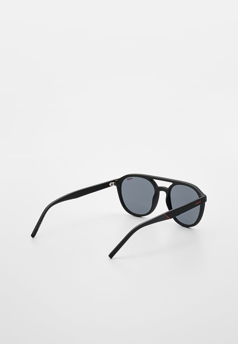 Schwarze Sonnenbrille mit runden Gläsern. Matt schwarze Fassung, schlankes Design und strukturierte schwarze Bügel, die mit einem roten Streifen akzentuiert sind.