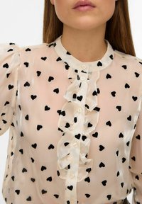 Prozirna, svijetlo roza bluza s crnim uzorcima srca, nabrana prednja strana i suptilno zatvaranje na dugmad. Kratke napuhane rukave dodaju volumen.