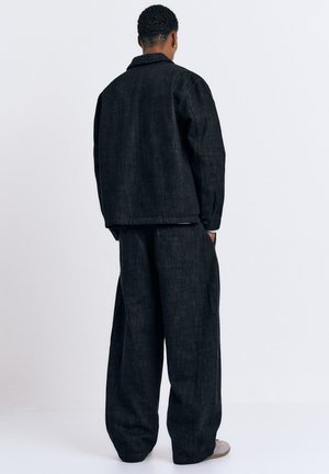 Mężczyzna stojący tyłem, ubrany w luźną czarną kurtkę jeansową i spodnie oraz beżowe buty, na jasnym, jednolitym tle.