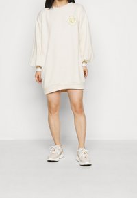 Kermanvärinen ylipitkä college-mekko, jossa on puffihihat ja pyöreä kaula-aukko. Rintataskussa on pieni brodeerattu logo.