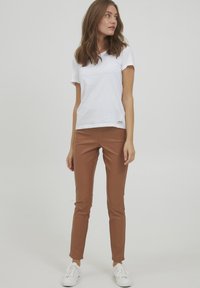 Witte katoenen t-shirt met een ronde hals en een klein logotag. Bruine faux leren skinny broek. Model staat in witte sneakers.