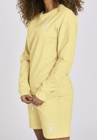Felpa gialla a maniche lunghe e pantaloni corti coordinati. Tessuto morbido con polsini a coste. Presenta un piccolo logo bianco sul petto e sui pantaloni.