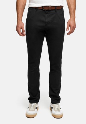 Homme portant un pantalon noir ajusté avec une ceinture marron, une chemise blanche et des baskets blanches avec des accents noirs et des semelles en gomme.