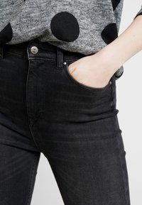 Jeans de denim negro con corte ajustado, que presentan un bolsillo frontal, cierre de botón plateado y una textura sutil. Combinados con una blusa gris con lunares negros.
