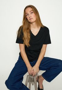 Jonge vrouw met lang lichtbruin haar, gekleed in een zwart V-hals T-shirt en blauwe jeans, zittend op een kruk tegen een effen achtergrond.