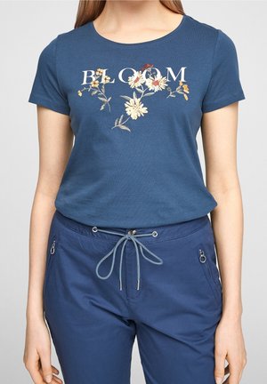 Frau trägt ein blaues T-Shirt mit dem Schriftzug "BLOOM" und floralem Muster, kombiniert mit marineblauen, mit Kordelzug und Reißverschlusstaschen ausgestatteten Hosen.