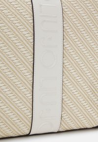 Bolso tejido beige con una correa de cuero blanco y un logo en relieve. La textura es suave con un patrón diagonal en el cuerpo.
