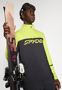 Svart og neongrønn halv-zip genser med "SPYDER"-logo, holdt med skistaver. Teksturert stoff, kontrastfargede ermer, sportsgoggles på hodet.