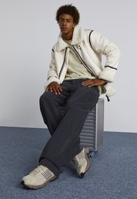 Vit fårskinnsjacka med svart trim, ljusgrön T-shirt, mörka jeans och beige sneakers. Modell sitter på en grå metallbehållare.