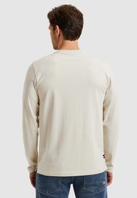 Langærmet cremefarvet skjorte lavet af blødt stof, med rund hals og sideslids, parret med blå jeans, der er synlige ved taljen.