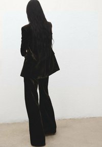 Čierny zamatový oblek s priliehavým blejzrom a rozšírenými nohavicami. Textília má jemnú štruktúru a mierny lesk, pozorovaná zozadu.