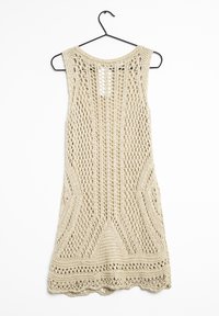 Robe sans manches en tricot de couleur beige clair, avec un motif ajouré, un ourlet festonné et un large décolleté, suspendue à un crochet noir.