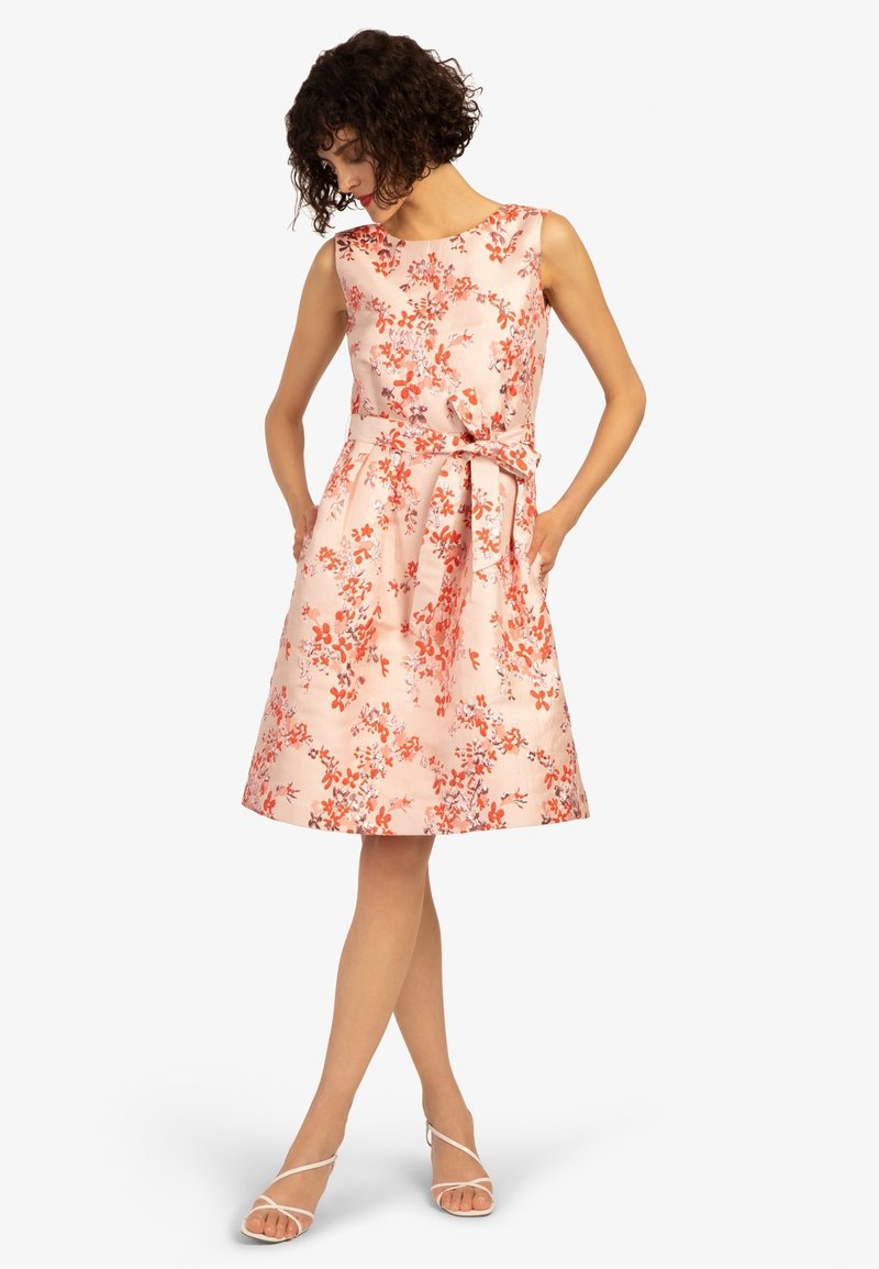 Femme debout, mains dans les poches, portant une robe sans manches rose pâle longueur genou avec un motif floral orange et une ceinture à nouer, talons blancs à lanières.