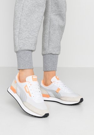 Person iført grå joggingbukser og hvide Puma-sneakers med fersken- og grå accenter, stående på et hvidt gulv mod en ensfarvet væg.