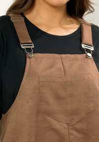 Pantalones de terciopelo marrón con clips de metal ajustables, que cuentan con un bolsillo frontal y una textura suave, usados sobre una camiseta negra, vista en picado.