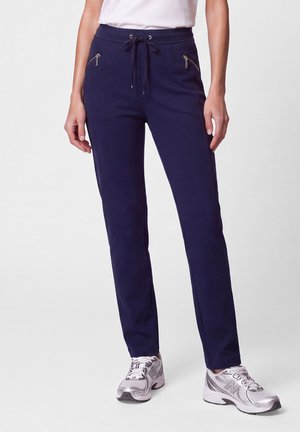 Woman standing wearing navy blue straight pants with zipper pockets and white sneakers, shown from waist to feet.