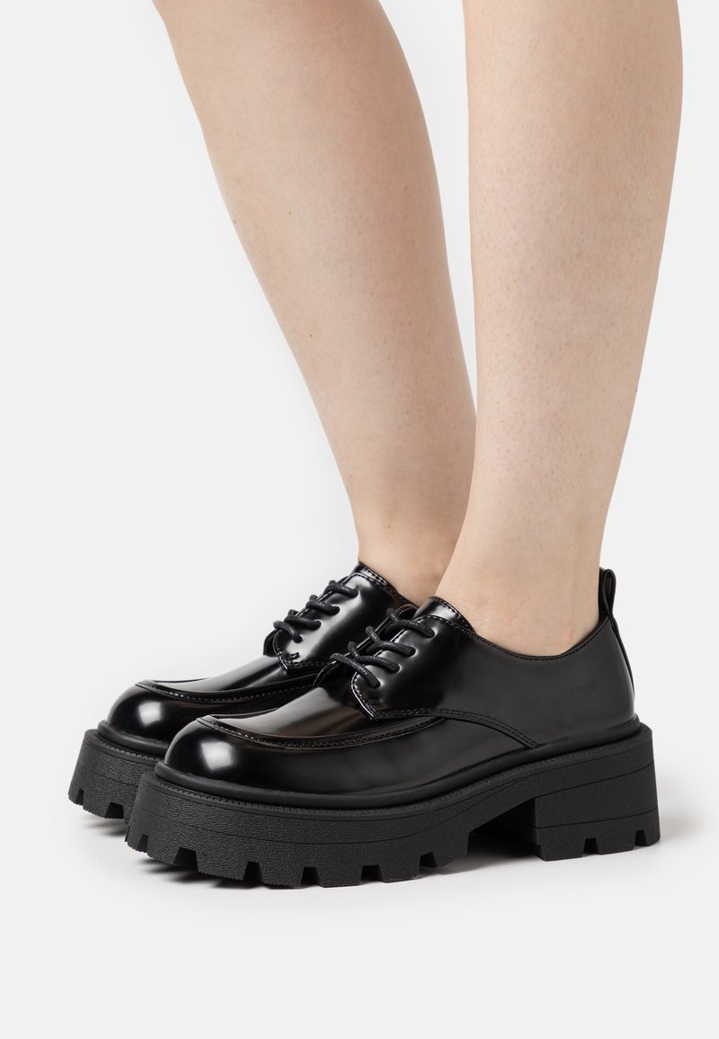 Chaussures en cuir verni noir avec une semelle épaisse et texturée noire. Elles disposent d'un bout rond et de six œillets pour lacets sur le dessus pour un maintien sécurisé.