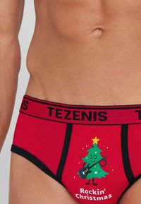 Czerwone bawełniane slipy z czarnym paskiem i napisem "TEZENIS", ozdobione grafiką rysunkowej choinki oraz tekstem "Rockin' Christmas."