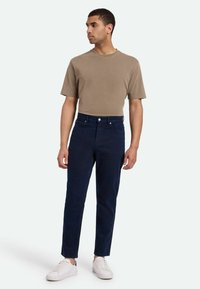 Camiseta de algodón marrón claro para hombre combinada con vaqueros de pierna recta azul marino. Zapatillas blancas completan el atuendo casual. Diseño sencillo, sin patrones.
