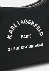 Sac en nylon noir avec du texte blanc en relief : "KARL LAGERFELD PARIS 21 RUE ST-GUILLAUME" et une fermeture éclair argentée en haut.