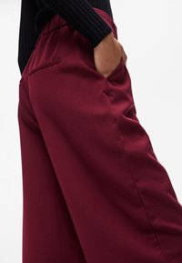 Burgundia laiad püksid siledast materjalist, elastse vöökoha ja küljetaskutega; omaduste seas on peened õmblusdetailid.