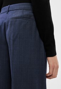 Persona, kas valkā tumši zilas piegrieztas bikses un melnu garu piedurkņu topu, roka atbalstīta pie sāniem pret gaišu fona.