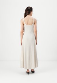 Lang, lys beige kjole med en tettsittende bodice, tynne stropper og en flytende skjørt. Glatt tekstur og minimalt design. Svarte hæl sandaler brukt.