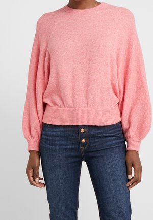 Femme portant un pull rose clair à manches longues avec taille et poignets côtelés, associé à un jean taille haute bleu foncé avec des boutons.
