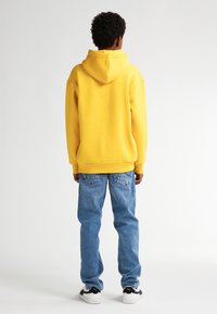 Felpa gialla con cappuccio, dalla vestibilità comoda, abbinata a jeans azzurri chiari e sneakers bianche e nere. La felpa ha una grande texture morbida.