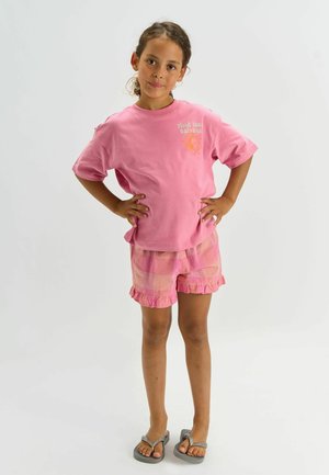 Vaaleanpunainen puuvillainen T-paita, jossa "Find Your Balance" -kuvio, yhdistetty vaaleanpunaisiin ja oransseihin ruutuhousuihin, joissa on röyhelöhelma, ja harmaat varvassandaalit.