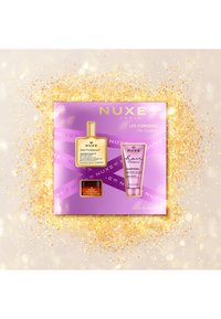 NUXE kosmetikset i en rosa låda innehåller en guldflaska, en röd burk och en rosa schamporör, alla märkta med tydlig text. Bakgrunden är prydd med guldglitter.