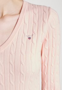 Maglione a collo a V in rosa chiaro con lavorazione a trecce. Design con scollo a V, logo discreto e piccolo emblema ricamato sul petto. Texture morbida.