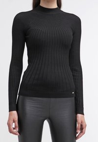 Pull noir en maille côtelée avec un col montant, coupe ajustée, manches longues, et un détail discret du logo à la taille, assorti à un pantalon noir.