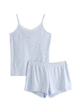 Młoda kobieta z kręconymi włosami, ubrana w niebiesko-biały, paskowany top bez rękawów i pasujące szorty, stojąca boso na jednolitym tle.